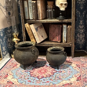May include: Two miniature black cauldrons with three legs each sit on a small rug with a blue and pink floral pattern. A bookshelf in the background holds miniature books, a skull, and a glass bottle.