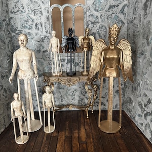 Op de afbeelding: Een verzameling engelenbeeldjes in verschillende maten en kleuren. De engelen omvatten een grote goudkleurige engel met gedetailleerde vleugels, een zwarte engel en verschillende wit- en goudkleurige figuren. De beeldjes worden tentoongesteld op een decoratieve tafel en een houten vloer.