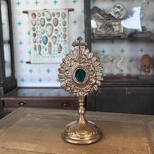 Puede incluir: Una reliquia religiosa dorada y ornamentada con una piedra verde en el centro, colocada sobre un soporte decorativo. El intrincado diseño presenta una cruz en la parte superior y adornos detallados. La reliquia se exhibe sobre una superficie de madera.
