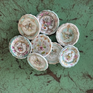 May include: A collection of small, antique porcelain plates with floral designs. The plates feature a variety of pastel colors, including pink, blue, and yellow, with scalloped edges. The plates are arranged on a green surface.