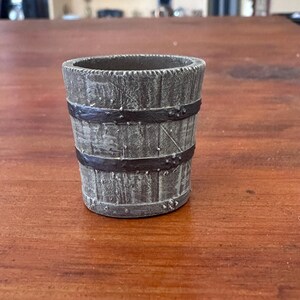 Puede incluir: Un pequeño cubo de madera de aspecto envejecido, con bandas metálicas oscuras. El cubo tiene una superficie texturizada, con bandas remachadas a la madera. Tiene forma cilíndrica y está sobre una superficie de madera.