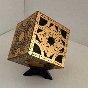 May include: A black wooden cube with gold painted symbols and a black star in the centre. The cube is on a black stand.