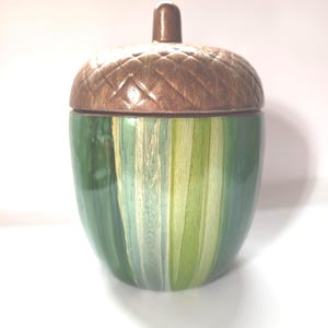 以下が含まれることがあります： 蓋付きの陶器製どんぐり型ジャー。蓋は茶色でテクスチャード加工が施されており、ジャーのベースはさまざまな色合いの垂直ストライプの緑色です。ジャーの上部には小さな茶色のノブが付いています。