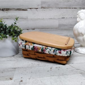 Può includere: Un cestino rettangolare in legno intrecciato con un coperchio in legno chiaro e una fodera in tessuto con un colorato motivo floreale. Il cestino è posizionato tra una piccola pianta verde in un vaso bianco e una statuetta di gallo in ceramica bianca.