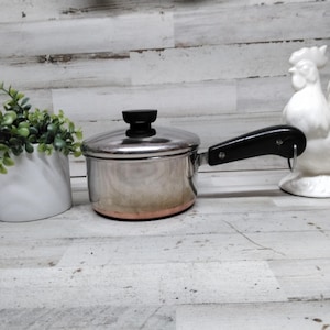 May include: A stainless steel saucepan with a black handle and lid, next to a white ceramic rooster figurine and a white pot with green foliage. The pot has a copper-colored bottom.