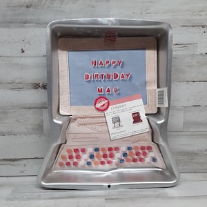 May include: A silver computer-shaped cake pan with a blue screen that says "Happy Birthday Mac". The pan is made by Wilton and has a red and white label on the side.