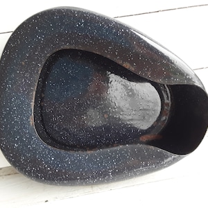 Puede incluir: Un orinal vintage azul marino con un patrón blanco moteado. El orinal de esmalte tiene una forma ovalada distintiva con un asa curva y una abertura ancha. La pieza antigua está sobre un fondo blanco.