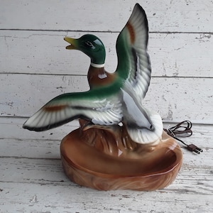 Puede incluir: Una lámpara de cerámica con forma de pato con un diseño pintado en verde, marrón y blanco. El pato está en posición de vuelo con las alas extendidas. La base de la lámpara es un plato de cerámica marrón con una superficie texturizada.