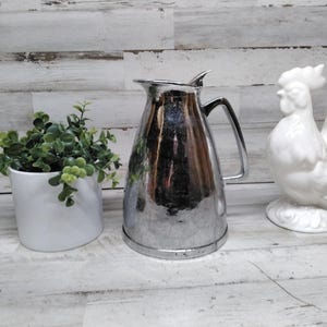 May include: A shiny, silver metal pitcher with a handle and a white ceramic rooster figurine. A small white pot with green leafy plants is also present. The items are arranged on a white wooden surface.