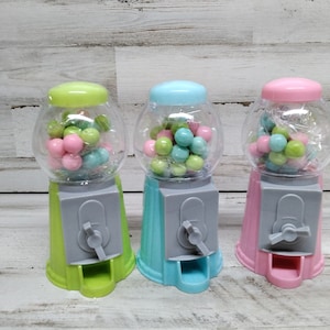 Może przedstawiać: Trzy miniaturowe automaty do gum do żucia, jeden zielony, jeden niebieski i jeden różowy, każdy wypełniony gumą do żucia w pastelowych kolorach.