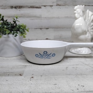 May include: White ceramic serving dish with a handle and blue floral design. A white ceramic rooster figurine and a small potted plant are in the background. The dish is likely vintage and suitable for serving food.