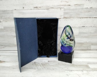 May include: A clear glass art piece with blue, green, and black swirls, set on a black base. The piece is next to an open, navy blue presentation box with a black velvet interior. The background is a white wood surface.