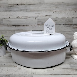 May include: An oval white enamel roasting pan with a speckled finish and a black rim. The pan features two side handles and a matching lid with a black handle. A small white house-shaped sign with the text "Vintage Tenn House" sits on the lid.