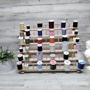 May include: A wooden thread spool organizer filled with various colored spools of thread. The spools are arranged on multiple levels, with a white ceramic rooster statue on the right side. A small potted plant is on the left.