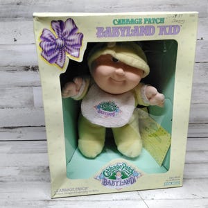 Peut inclure: Une poupée Cabbage Patch Kid Babyland Kid vêtue d'une tenue jaune et blanche avec un bavoir qui dit "Cabbage Patch Babyland". La poupée est dans une boîte avec un fond vert et blanc et un nœud violet.