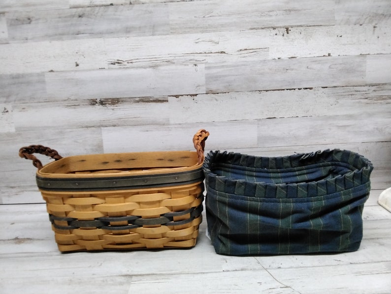 May include: Two storage baskets. The first is a rectangular woven basket with a light brown and grey design, brown braided handles, and a dark grey trim. The second is a blue and green plaid fabric basket.