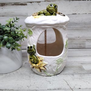 May include: A white ceramic frog-themed decorative container. The container has a large opening and is adorned with green frog figurines and lily pads. A gold-colored chain hangs inside. A potted plant is to the left.