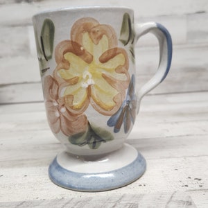 May include: A white ceramic mug with a floral design. The mug has a handle and a pedestal base. The flowers are painted in shades of yellow, orange, and brown.