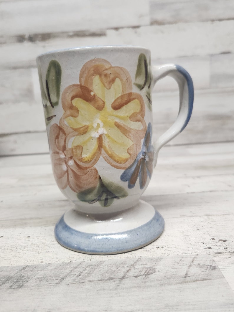 May include: A white ceramic mug with a floral design. The mug has a handle and a pedestal base. The flowers are painted in shades of yellow, orange, and brown.