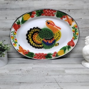 May include: Oval white enamelware platter with a colorful rooster design. The platter has a black rim and is decorated with fruits and vegetables. A white ceramic rooster figurine is on the right.