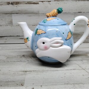May include: A blue and white ceramic teapot with a white rabbit design and a carrot on the lid. The teapot is decorated with carrots and has a blue and white glaze.