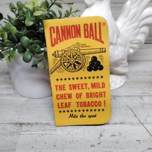 Puede incluir: Un paquete de tabaco Cannon Ball amarillo con texto rojo y una ilustración de cañón. El texto dice: "THE SWEET, MILD CHEW OF BRIGHT LEAF TOBACCO!" y "Hits the spot."