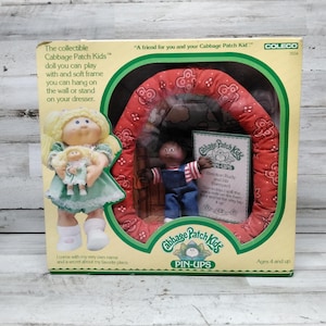 May include: A vintage Cabbage Patch Kids Pin-Ups doll set in its original packaging. The box features a doll holding a smaller doll, and a second doll inside a red bandana-covered frame. The box has text and is suitable for ages 4 and up.