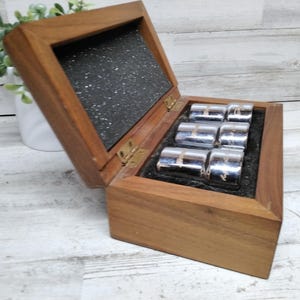 Peut inclure: Une boîte en bois avec un couvercle à charnière, ouverte pour révéler six objets argentés. La boîte est en bois foncé et doublée de tissu noir. Les objets argentés sont cylindriques et semblent décoratifs.