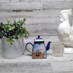 May include: A decorative teapot with a coastal scene, featuring a blue handle and spout, and a white ceramic rooster figurine. A white ceramic pot with green foliage is to the left of the teapot.