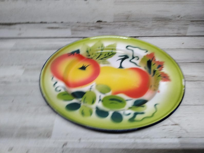 Peut inclure: Une assiette en &eacute;mail vintage avec un bord vert et un centre blanc. L'assiette pr&eacute;sente un motif peint &agrave; la main de pommes rouges et jaunes avec des feuilles vertes.