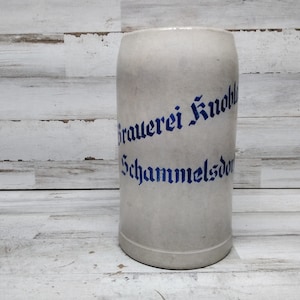 May include: An off-white ceramic beer stein with blue lettering, reading "Brauerei Knobloch Schammelsdorf". The stein has a cylindrical shape with a rounded base. The background is light-coloured, weathered wood.