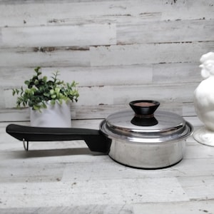 Puede incluir: Una cacerola de acero inoxidable con asa y tapa negras, con un pomo marrón oscuro. Una figura de gallo de cerámica blanca y una pequeña planta en maceta están en el fondo. La cacerola está diseñada para cocinar.