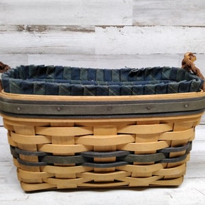 May include: Woven basket with a light brown and dark green pattern. The basket has a dark grey rim and brown braided handles. The interior is lined with a blue and green plaid fabric. A decorative storage container.
