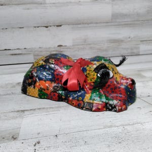 Könnte beinhalten: Eine farbenfrohe Patchwork-Hundefigur aus Stoff mit einer roten Schleife. Der Hund liegt mit dem Kopf zur Seite gedreht. Die Figur ist aus Papiermaché gefertigt und hat eine glänzende Oberfläche.