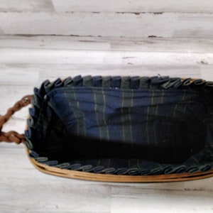 May include: A rectangular wicker basket with braided leather handles. The interior is lined with a blue and green plaid fabric. The rim of the basket is trimmed with a dark blue fabric. The basket is empty.
