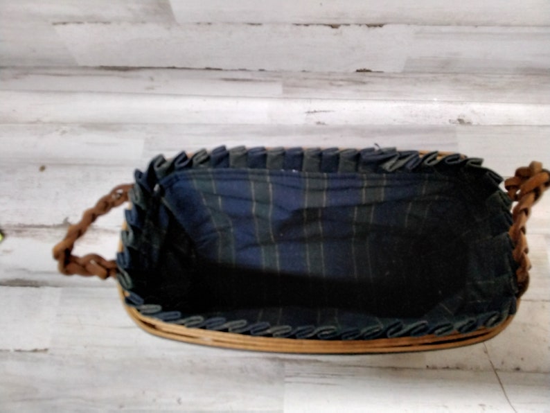 May include: A rectangular wicker basket with braided leather handles. The interior is lined with a blue and green plaid fabric. The rim of the basket is trimmed with a dark blue fabric. The basket is empty.