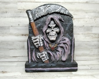 May include: A Halloween-themed decorative piece featuring a grim reaper figure. The reaper is depicted with a skeletal face, wearing a hooded robe, and holding a scythe. The color palette includes shades of gray, black, and purple.