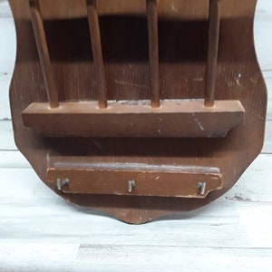 May include: A vintage wooden wall-mounted key and mail holder. The shield-shaped holder is made of dark brown wood and features a shelf, four vertical dowels, and three key hooks. The wood has visible wear and tear.