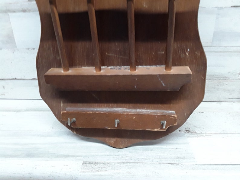 May include: A vintage wooden wall-mounted key and mail holder. The shield-shaped holder is made of dark brown wood and features a shelf, four vertical dowels, and three key hooks. The wood has visible wear and tear.