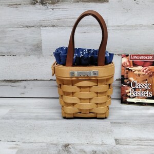 May include: A small, brown wicker basket with a leather handle and a blue and white patterned fabric lining. The basket is sitting on a light wood surface next to a small book titled "Longaberger Classic Baskets".