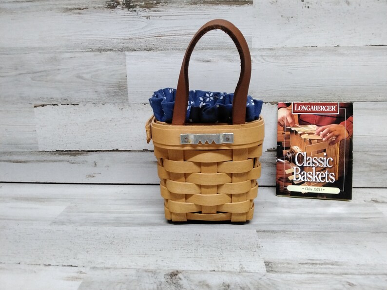 May include: A small, brown wicker basket with a leather handle and a blue and white patterned fabric lining. The basket is sitting on a light wood surface next to a small book titled "Longaberger Classic Baskets".