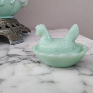 Puede incluir: Una figura de gallina sobre nido de vidrio verde claro. La gallina está sentada sobre una base en forma de cuenco a juego. La figura está hecha de un material de vidrio translúcido. La gallina tiene plumas detalladas y un pico definido. La base tiene un borde festoneado.