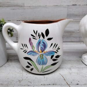 Puede incluir: Jarra de cerámica blanca con borde marrón y asa. La jarra está decorada con un diseño de flor de iris azul y morado, rodeada de hojas negras. Un gallina de cerámica blanca está en el fondo.