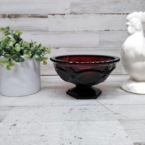 Pode incluir: Uma natureza morta com uma tigela de vidro vermelho rubi com um padrão decorativo, uma figura de galo de cerâmica branca e uma floreira de cerâmica branca com folhagem verde. A tigela está sobre um pedestal preto.
