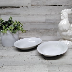 May include: A still life composition featuring two small, oval white ceramic bowls, a white ceramic rooster figurine, and a white cylindrical planter with green foliage. The items are on a light-coloured, weathered wooden surface.