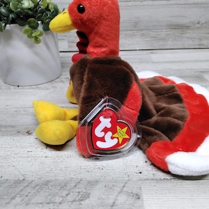 May include: A plush turkey toy with a red head and wattle, brown body, and a tail with red, white, and brown sections. The turkey has yellow feet and a yellow beak. A red tag is attached to the toy.