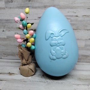 May include: A large, light blue plastic Easter egg with a bunny rabbit design. The egg is sitting on a wooden surface next to a small burlap bag filled with colorful Easter eggs.