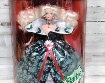 May include: A Special Edition Barbie doll in a red and clear display case. The doll has blonde hair, a white collar with holly accents, and a teal and black gown. The case reads "Happy Holidays" and "Barbie".