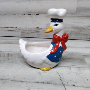 May include: A whimsical ceramic planter shaped like a goose wearing a chef's hat and apron. The goose is white with a yellow beak and feet, a red bow tie, and a blue apron with white polka dots. The planter has an open bowl for holding plants.