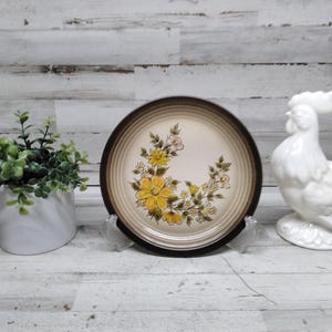 Puede incluir: Un plato decorativo con un diseño floral con flores amarillas y blancas, colocado sobre un soporte transparente. También se ve una figura de gallo de cerámica blanca y una pequeña planta en maceta.